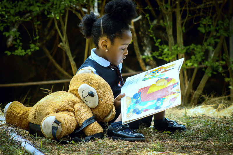 Young girl in pinifore sits on grass beside trees with a large teddybear reading a picture book